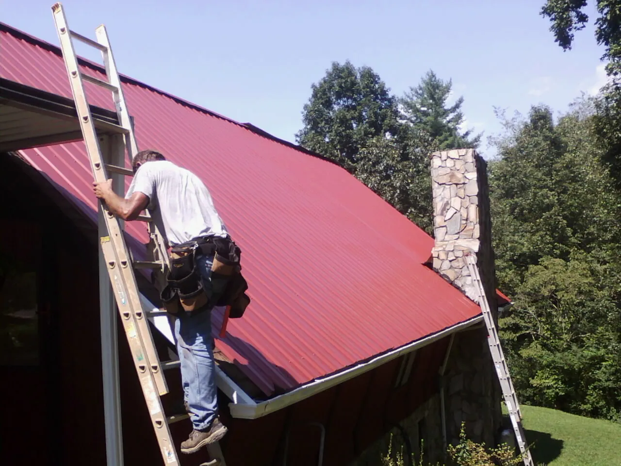 Roofer on a ladder beside a red metal roof and stone chimney — Soffit & Fascia Repair by Roofing Brothers in Delia